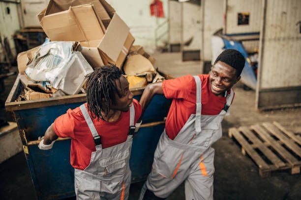 Two workers in overalls struggle to push a full dumpster, smiling.