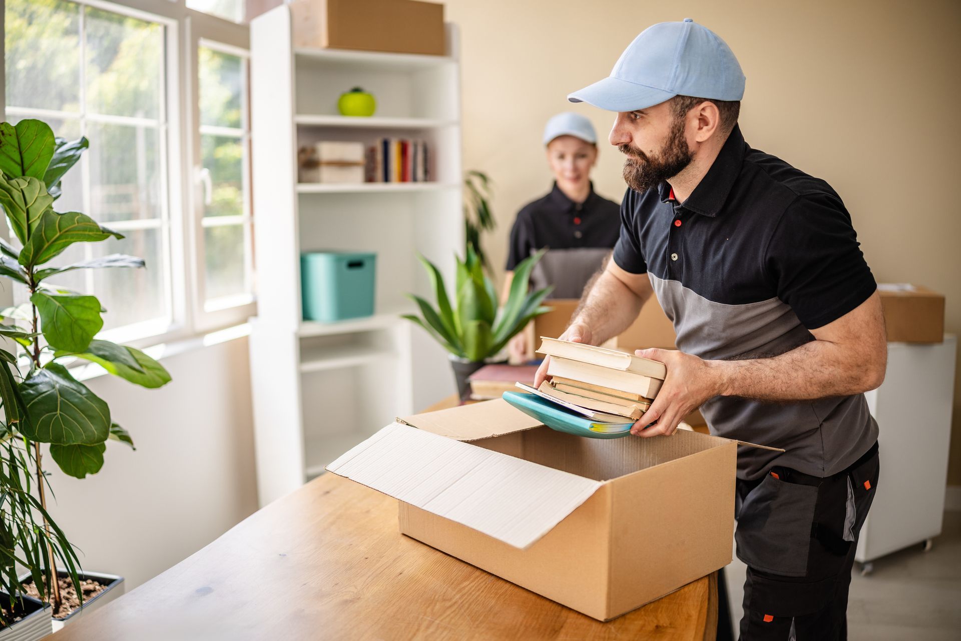 Two movers packing books and a tablet into a cardboard box in a room.