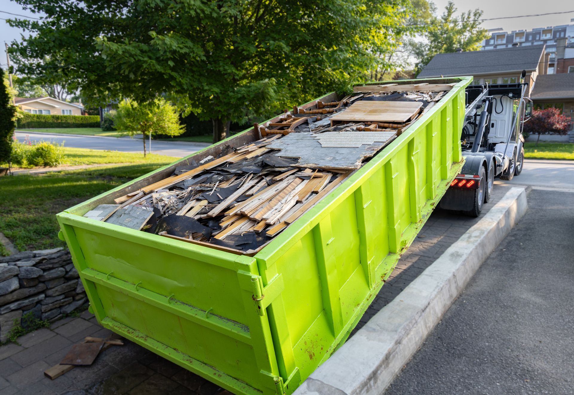 Green metal dumpster on a sidewalk under a blue canopy, with a white bag nearby.