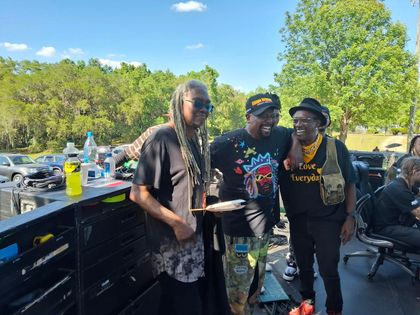 Three people posing beside a picnic table in a sunny park with trees and drinks nearby