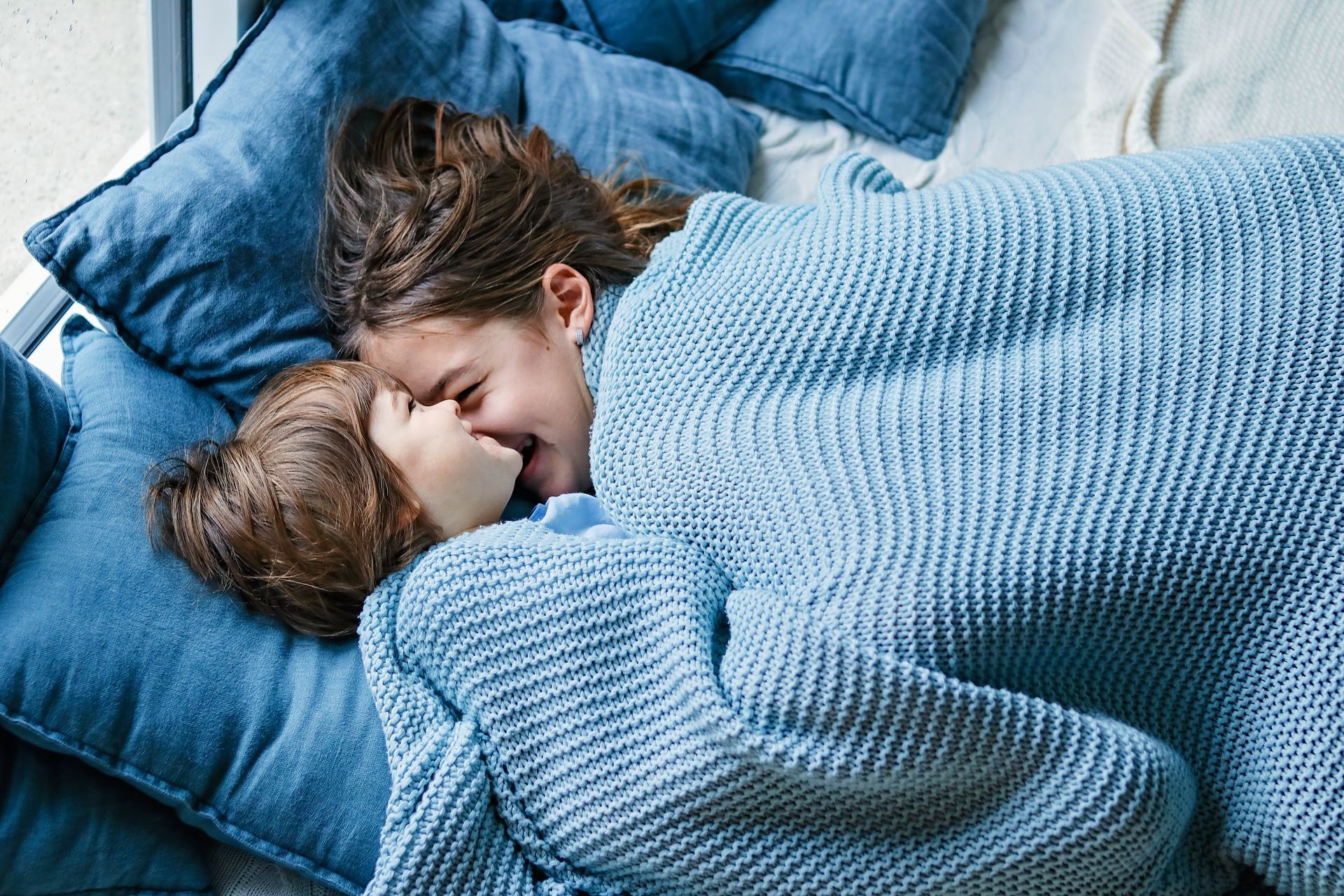 A woman and a child are laying under a blue blanket on a bed.