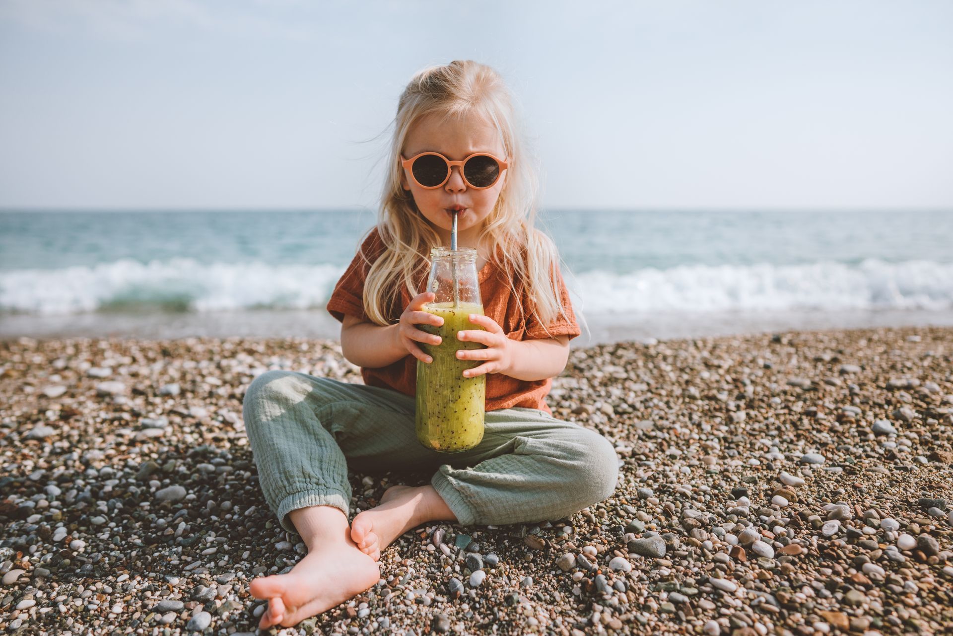 A little girl is sitting on the beach drinking a smoothie through a straw.