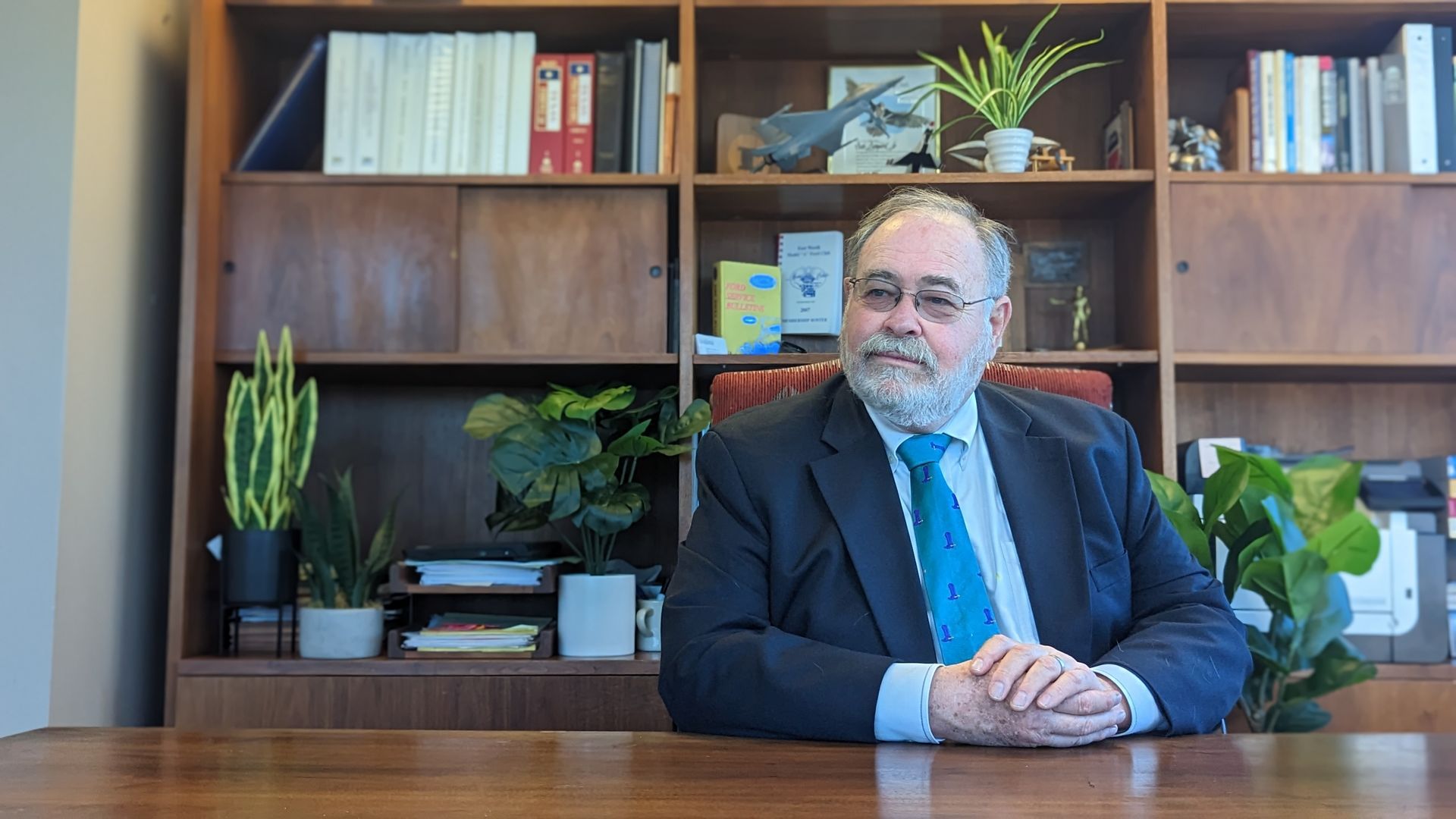 A man in a suit and tie is sitting at a desk in front of a bookshelf.