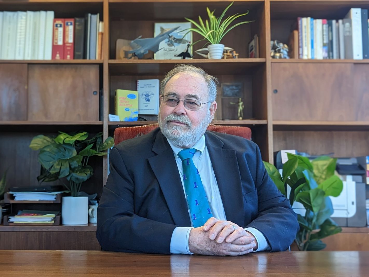 A man in a suit and tie is sitting at a desk in front of a bookshelf.