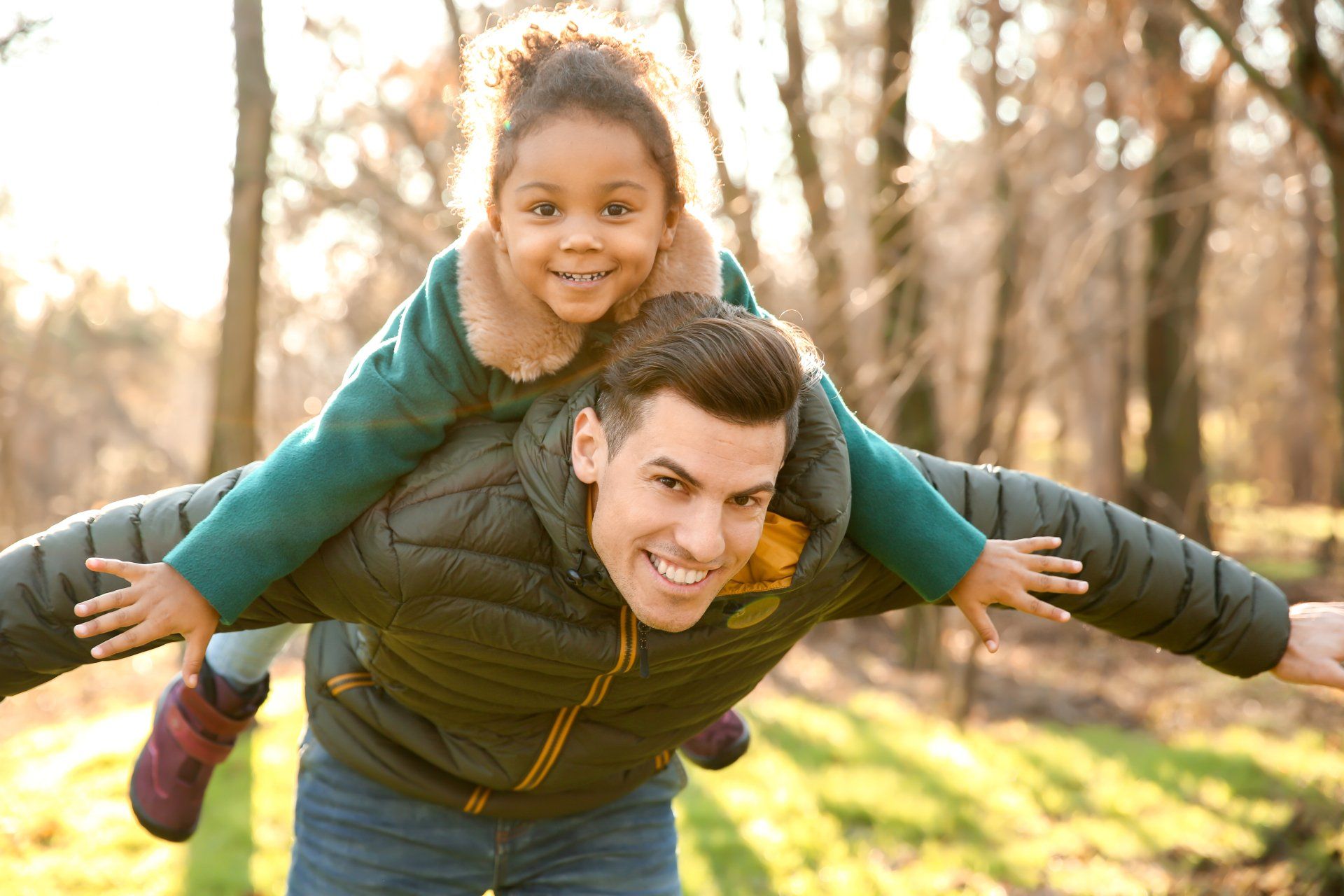 A man is carrying a little girl on his shoulders in the park.