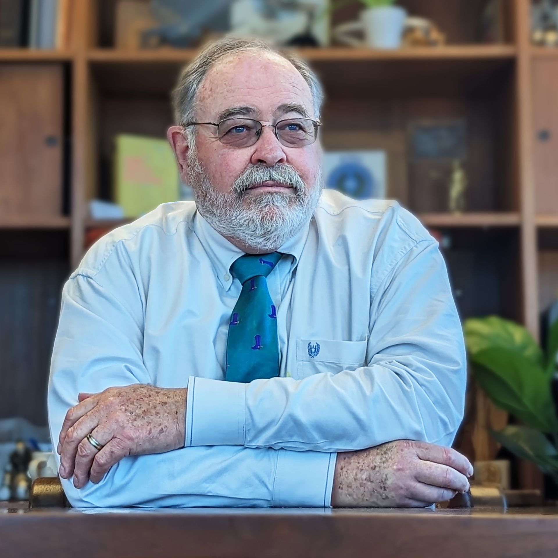 A man with a beard and glasses is sitting at a desk with his arms crossed.
