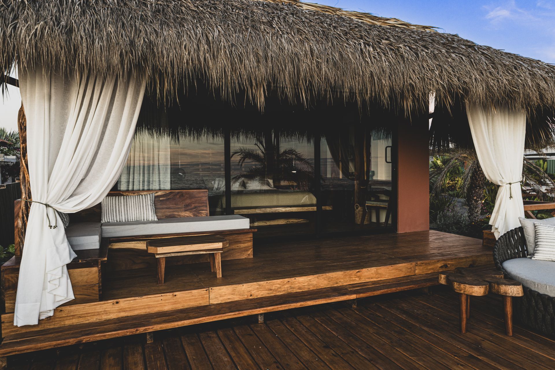 Una choza con techo de paja, terraza de madera y cortinas blancas.