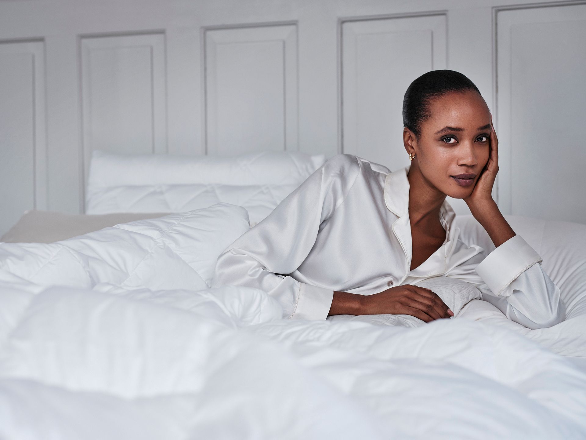 Woman in white shirt reclines on white bed, resting her head on her hand.