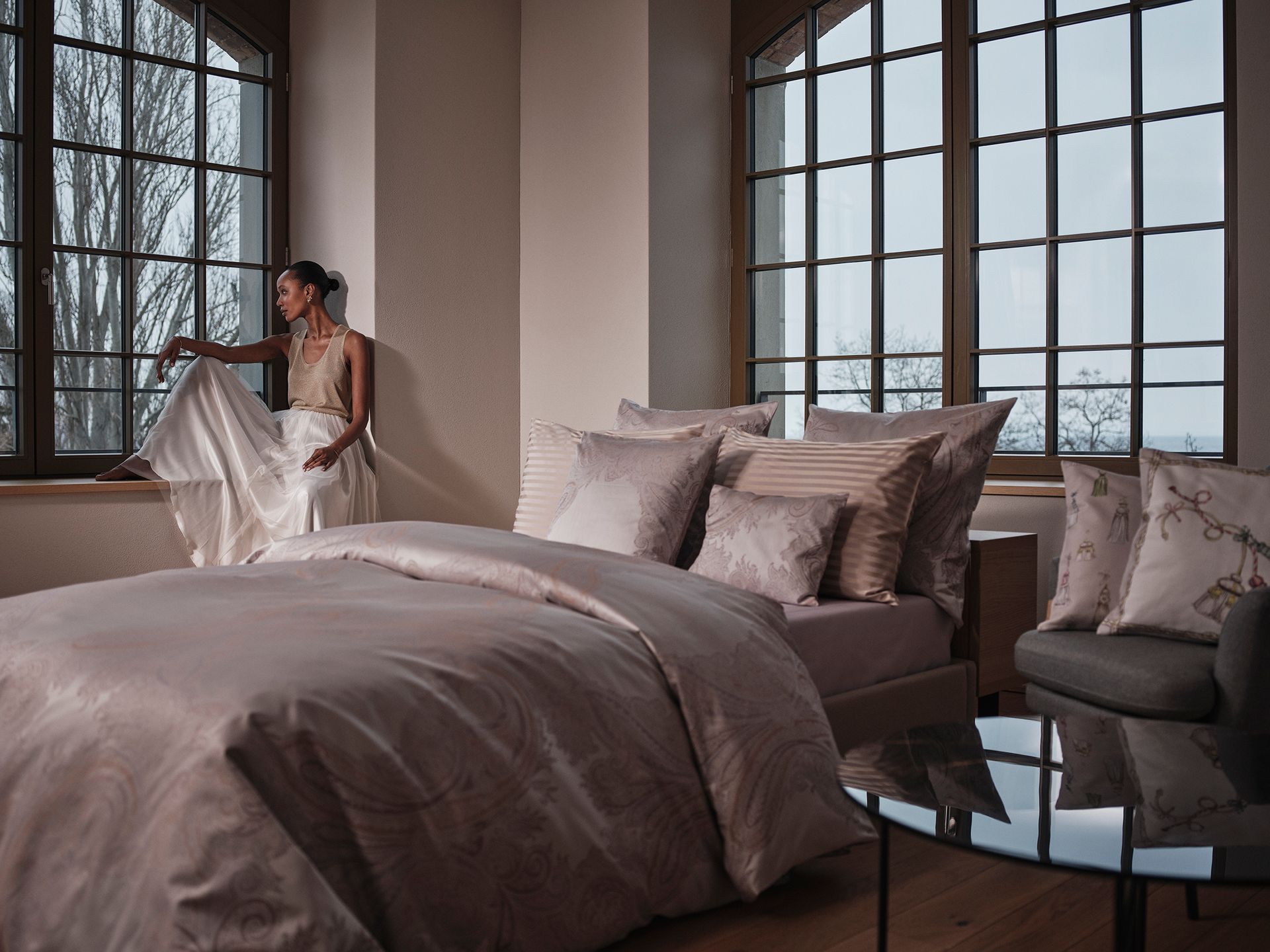 A person in a light, flowing gown sits by a large window in a bedroom with taupe bedding and soft, neutral decor.