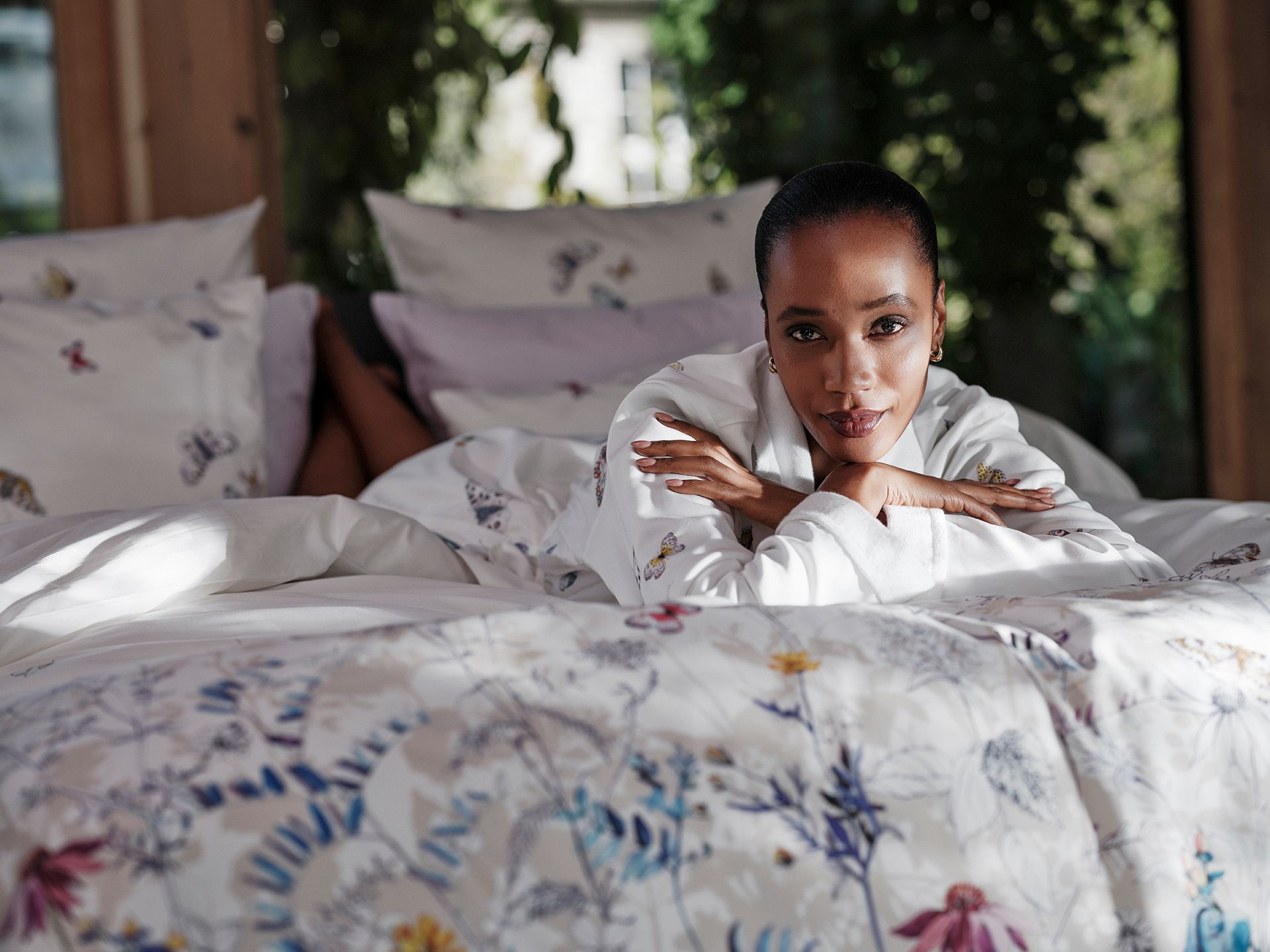 Woman in white robe, lying on bed with floral bedding, looking at the camera. Soft sunlight.