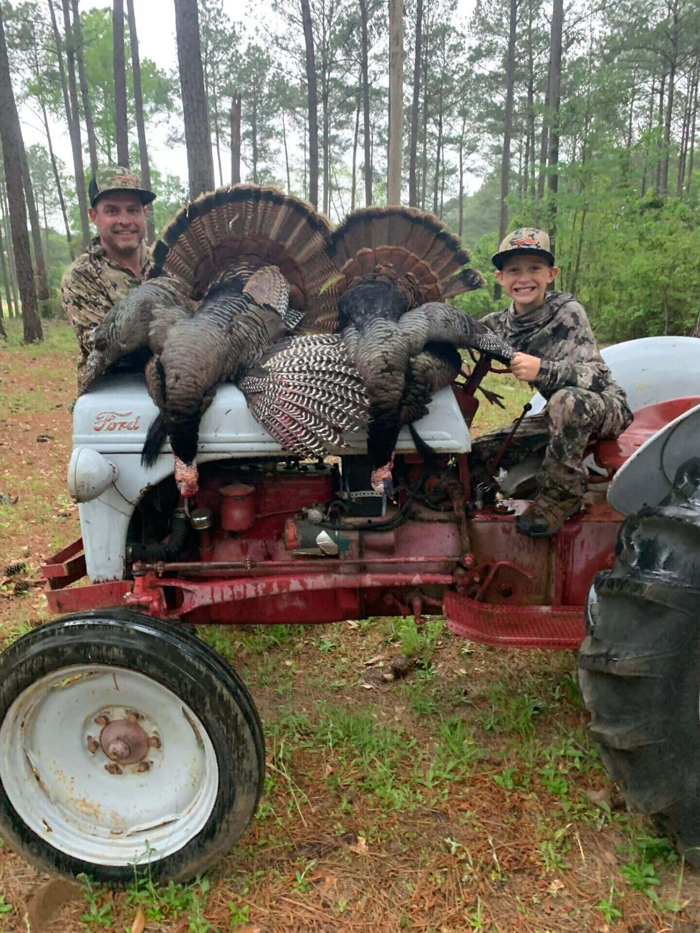 Two hunters posing with two wild turkeys on a red tractor in a wooded area