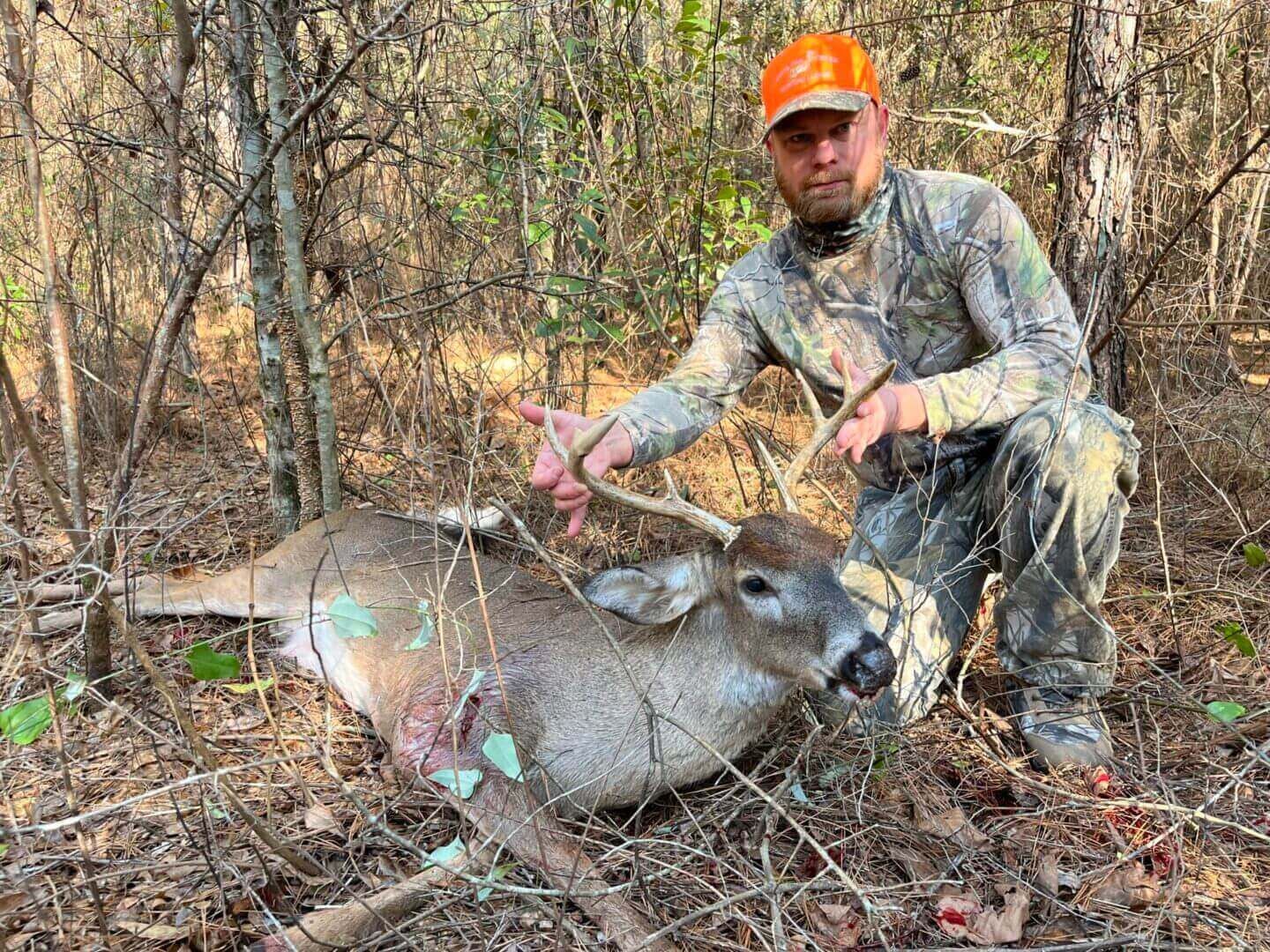 Hunter in orange cap kneels beside a downed deer in a wooded area.