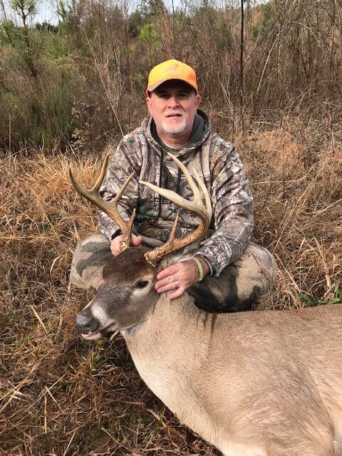 Hunter kneeling beside a large deer in brush, wearing camouflage and a yellow cap.