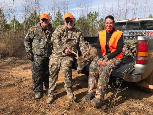 Three hunters posing by a pickup truck with a deer in a wooded field.