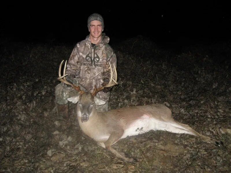 Hunter kneels beside a deer at night in a grassy field, holding it by the antlers.