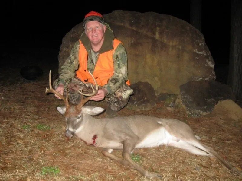 Hunter kneeling beside a deer on grass at night, wearing orange vest in front of a large rock