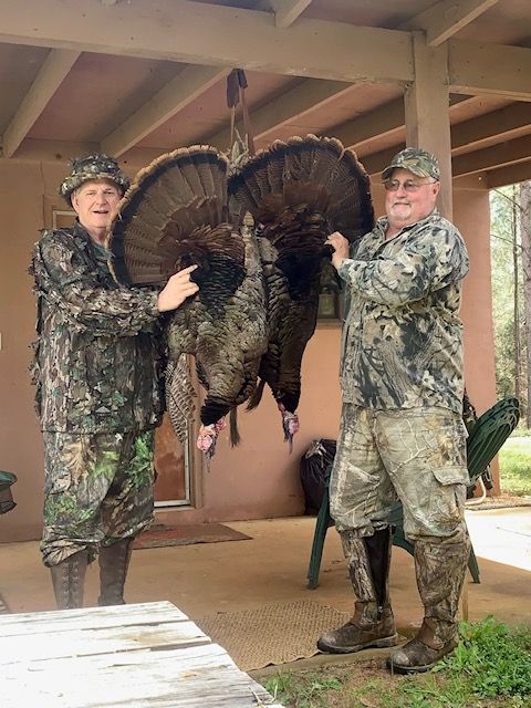 Two hunters holding a large wild turkey by the tail under a covered porch.