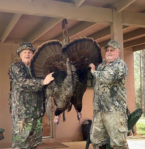 Two camo-clad hunters hold a large wild turkey by the tail on a porch.