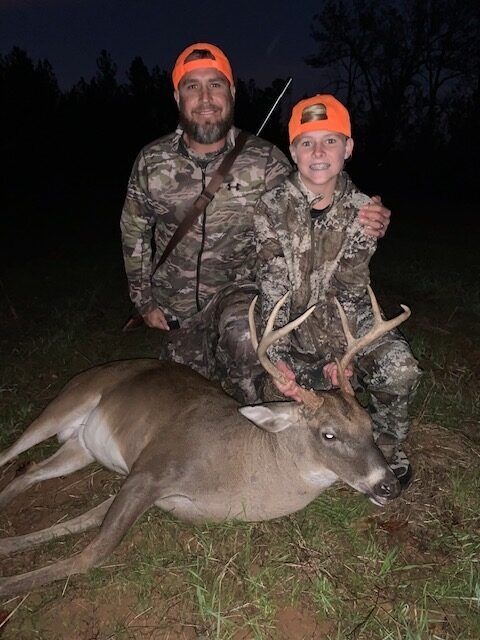 Two hunters in camo with orange caps pose over a deer at night.