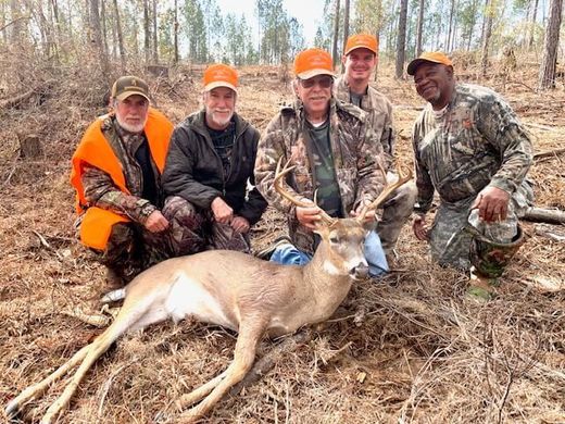 Five hunters kneel behind a deer in a grassy field, wearing orange hats and camouflage.