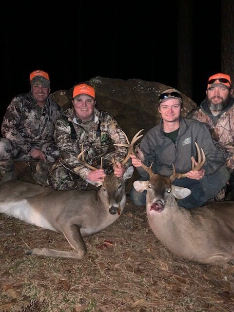 Four hunters pose at night with two deer and rifles in a wooded area.