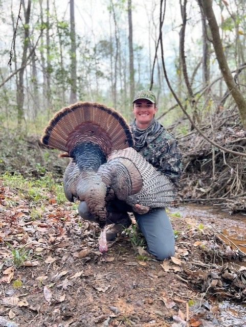 Hunter kneeling beside a large wild turkey in a leaf-strewn forest.