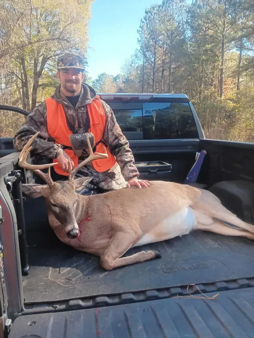 Hunter in orange vest poses with a deer in a pickup truck bed in the woods.