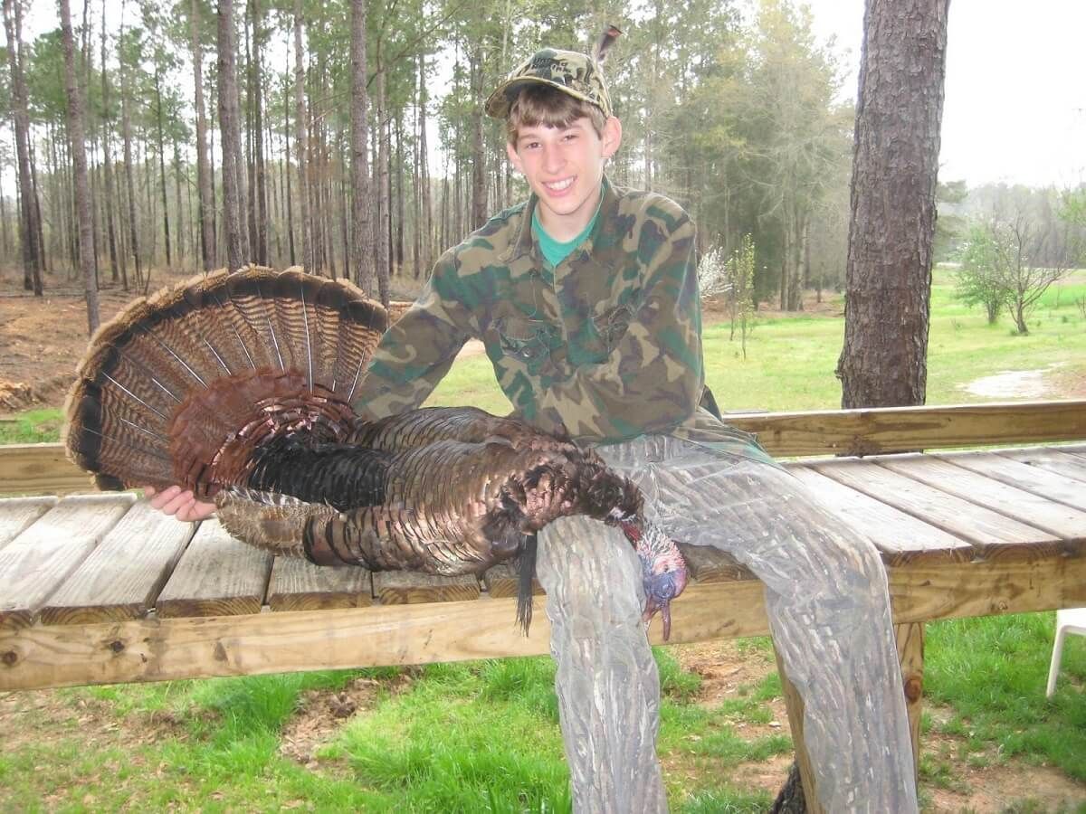 Person in camouflage posing with a wild turkey on a wooden bench outdoors