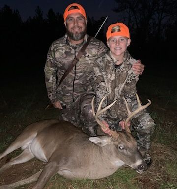 Two hunters in camo and orange hats pose with a deer at night.