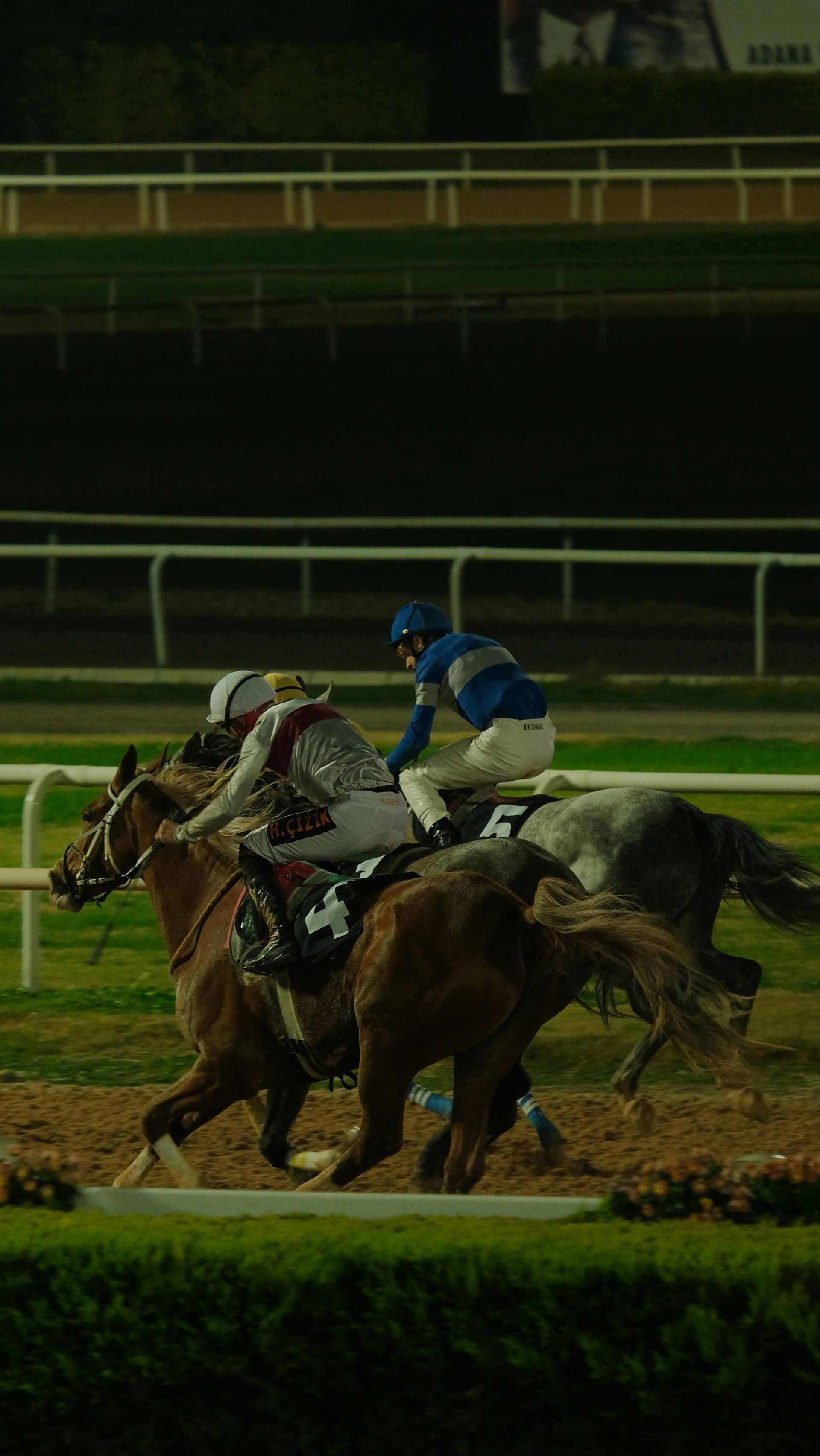 Two jockeys racing horses on a dirt track at night, one in blue and one in white silks