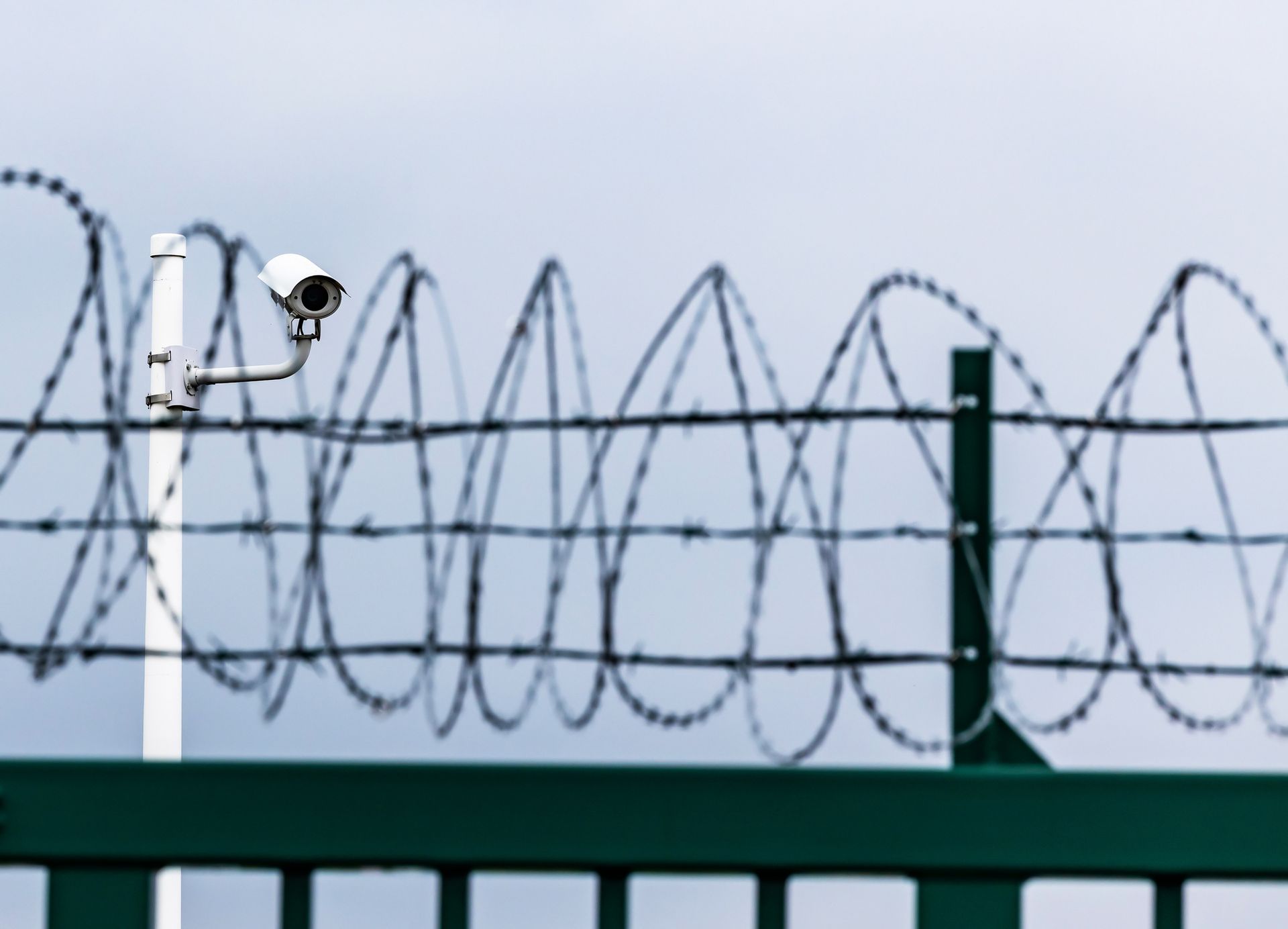 Security camera on a white pole, overlooking a green fence topped with barbed wire, under a cloudy sky.
