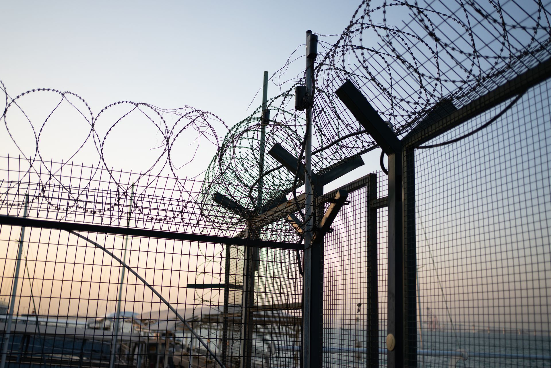 Razor wire atop a metal fence, silhouetted against a dusky sky.