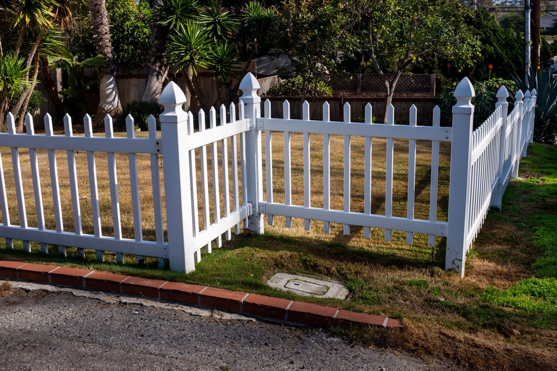 White picket fence enclosing a small grassy area; brick edging and trees in the background.