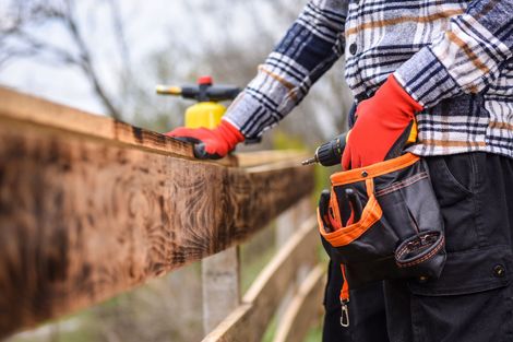 Person in a plaid shirt, red gloves, and tool belt using a drill on a wooden fence.