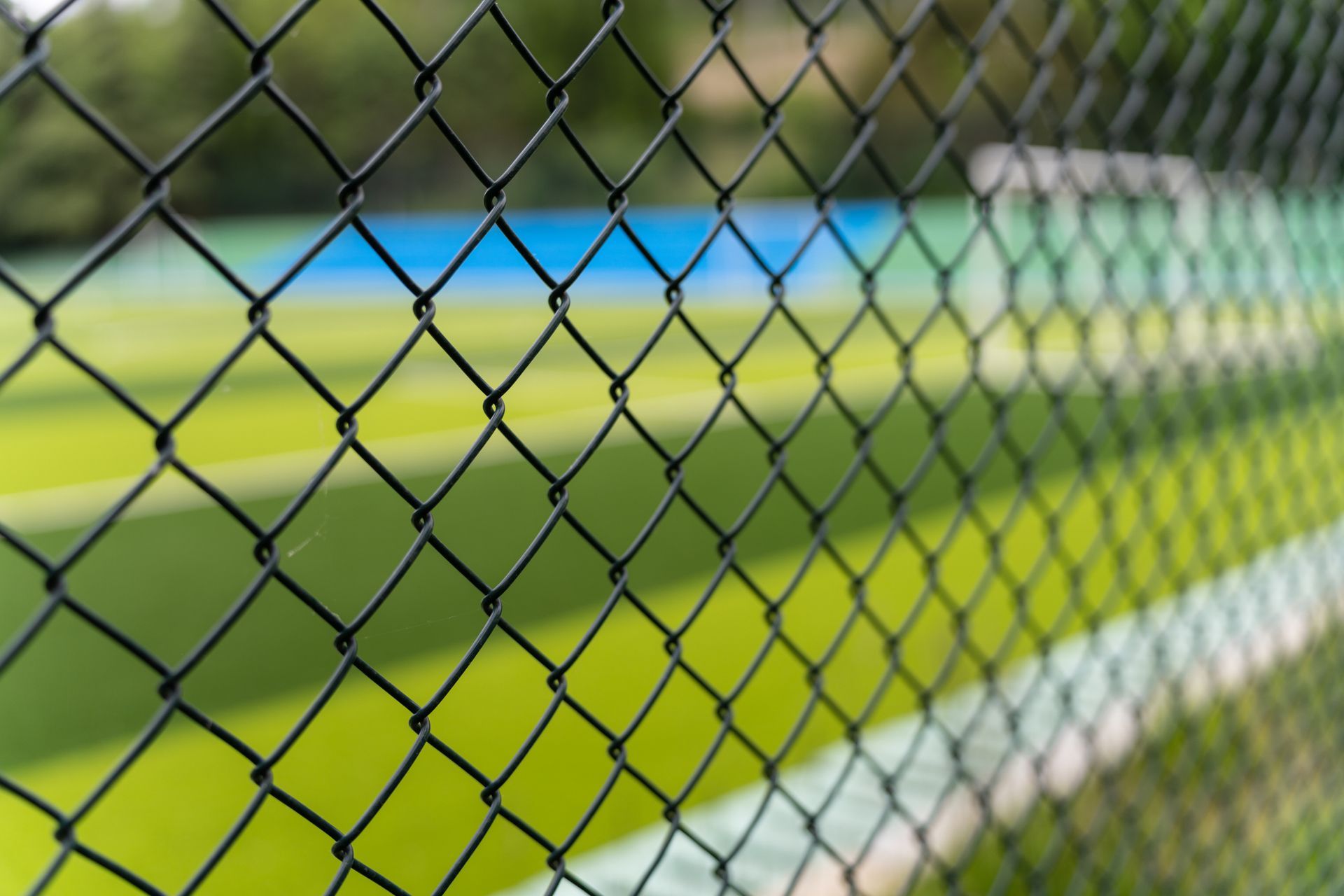 Black chain-link fence in the foreground with a tennis court in the background. Green and blue colors. Black chain-link fence in the foreground with a tennis court in the background. Green and blue colors.
