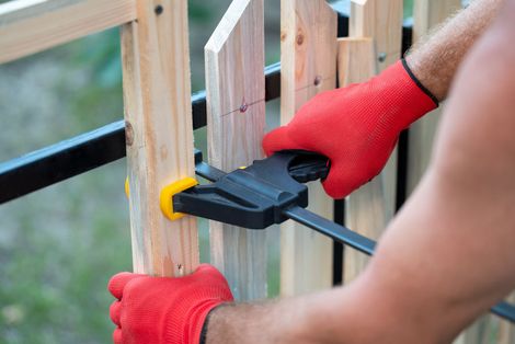 Hands in red gloves using a clamp to secure wooden fence slats.