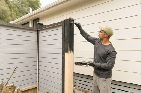 Man in gray sweater and cap painting a fence post black outside a house.