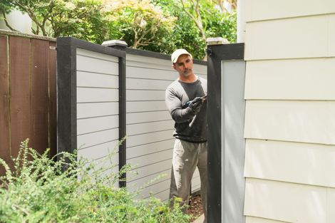 Man painting fence with black trim, wearing work clothes, outside, near house.