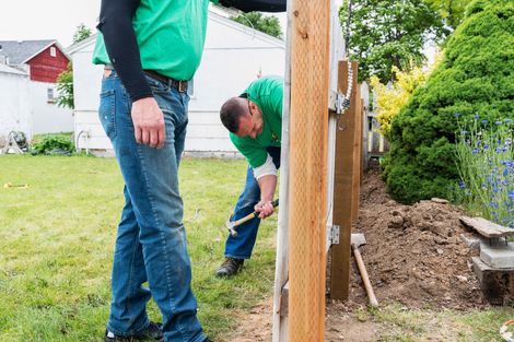 Two people building a wooden fence outdoors; one hammers, the other steadies the post.