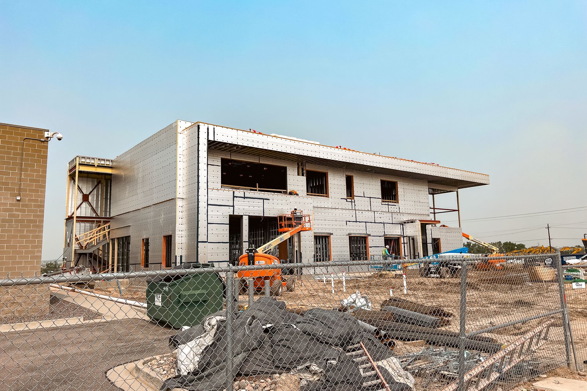 A large building is being built in the middle of a dirt field behind a chain link fence.