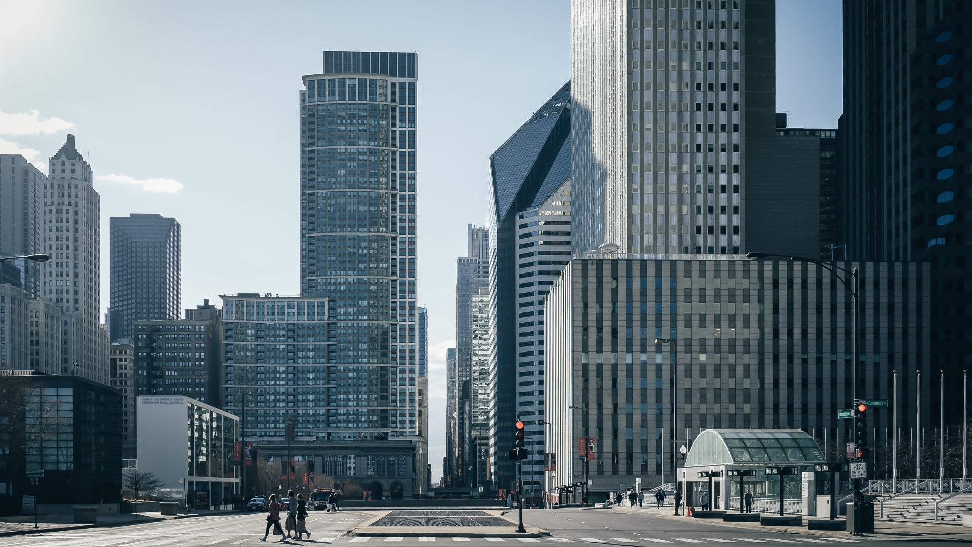 Skyscrapers line a city street under a bright, clear sky; people walk along the sidewalks.