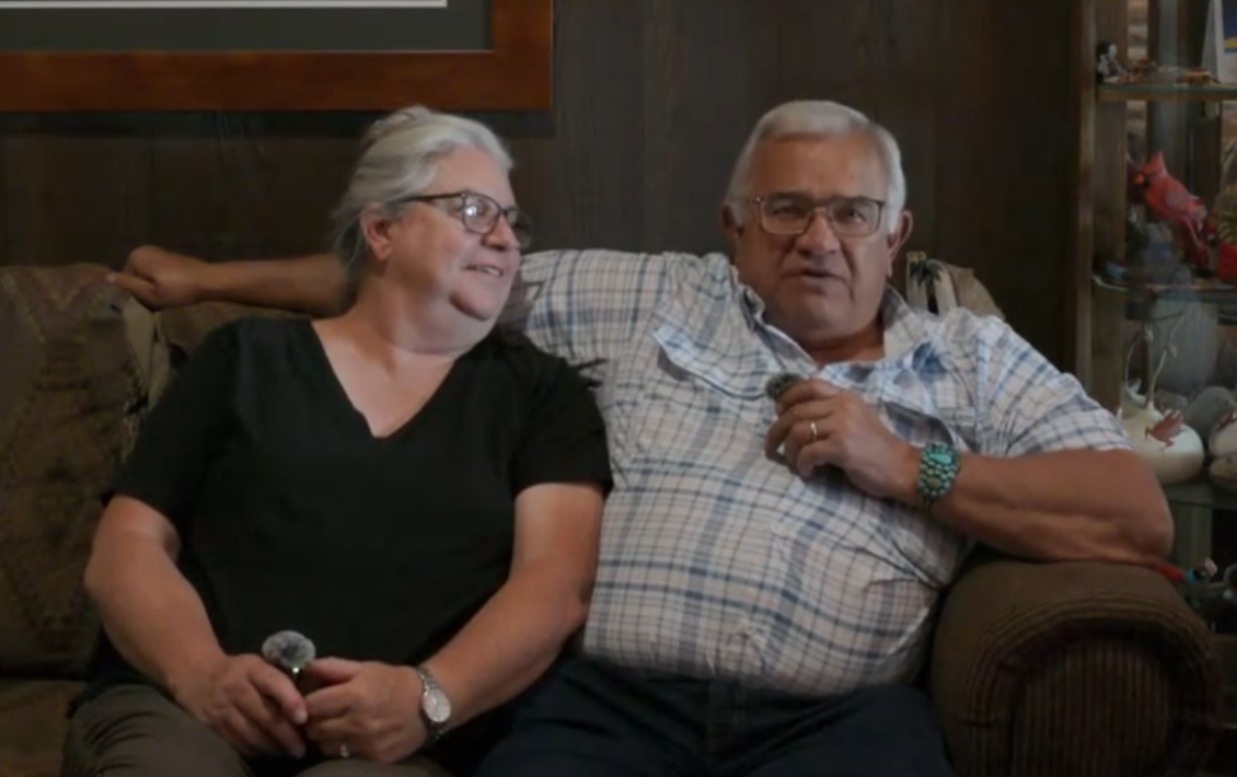 Couple sitting on a couch. The man has an arm around the woman. Both wear glasses.