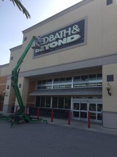 Bed Bath & Beyond store sign being worked on by a person in a lift, tan building.