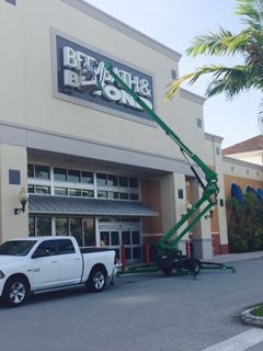 A green aerial lift near a Bed Bath & Beyond store, next to a white pickup truck.