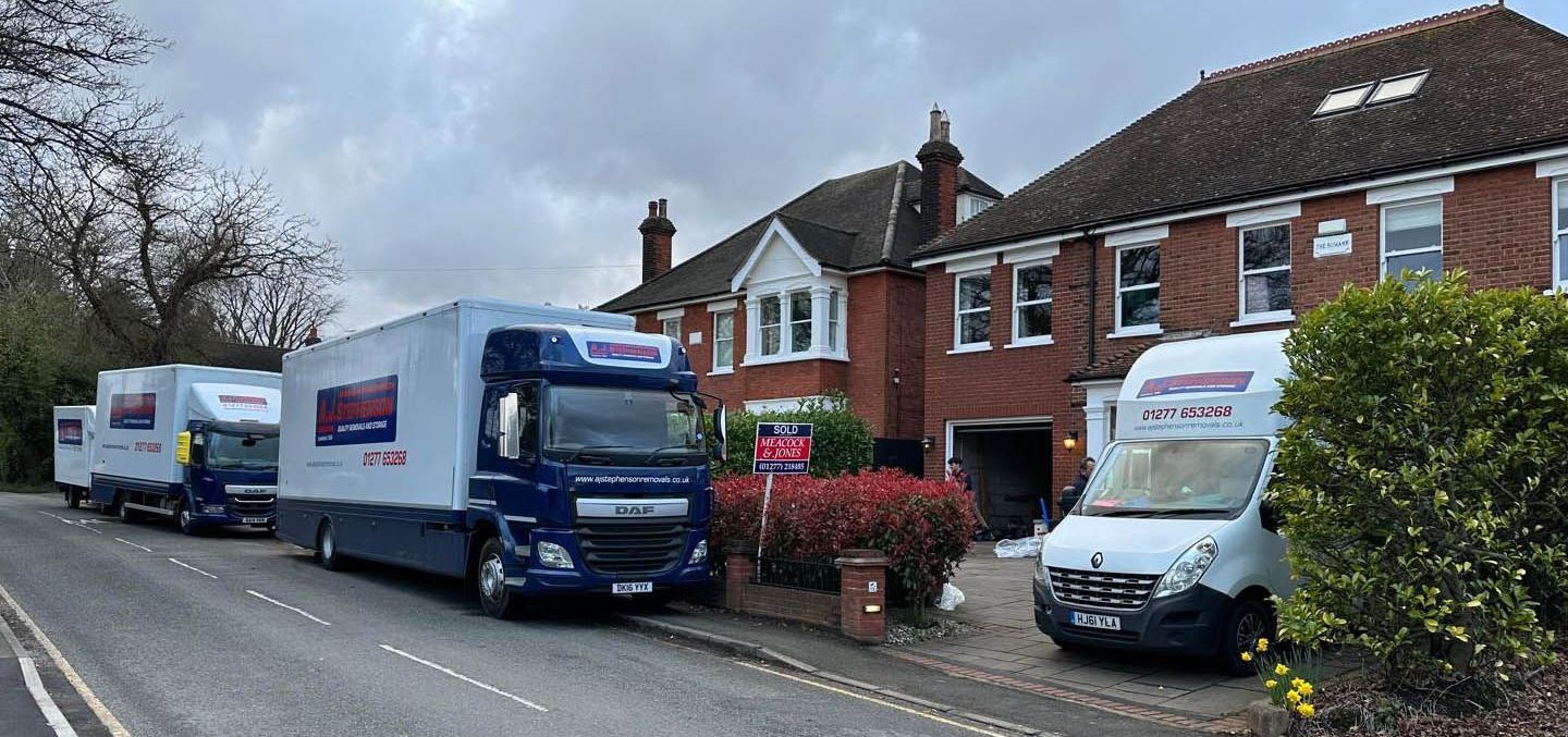 A row of AJ Stephenson Removals moving lorries are parked on the side of the road in front of a brick house.