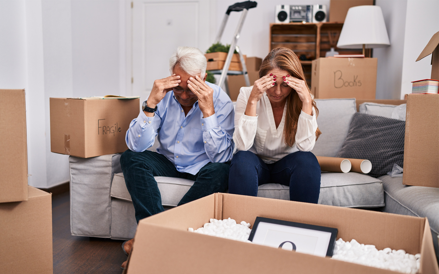 Two people on a couch cover their faces amid moving boxes in a packed living room.
