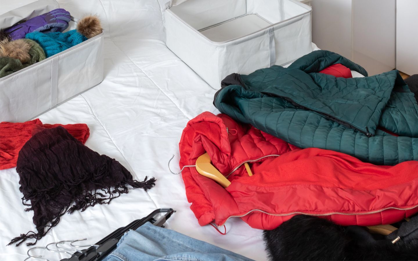 Clothes scattered on a white bed; red and green jackets, black scarves and a basket of items.