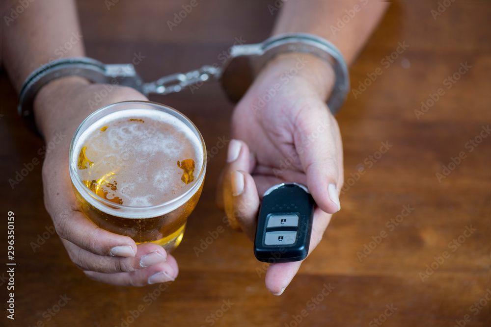 A close-up of a glass of alcohol and car keys. In the background, a man is in handcuffs.