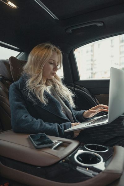 A woman is sitting in the back seat of a car using a laptop computer.