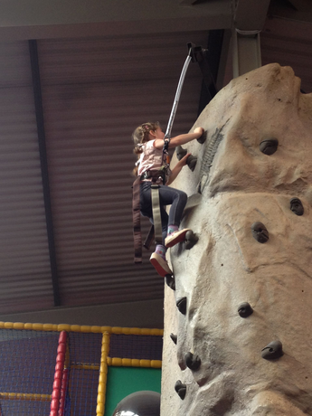 Girl in harness climbing an indoor rock wall, reaching for handholds.